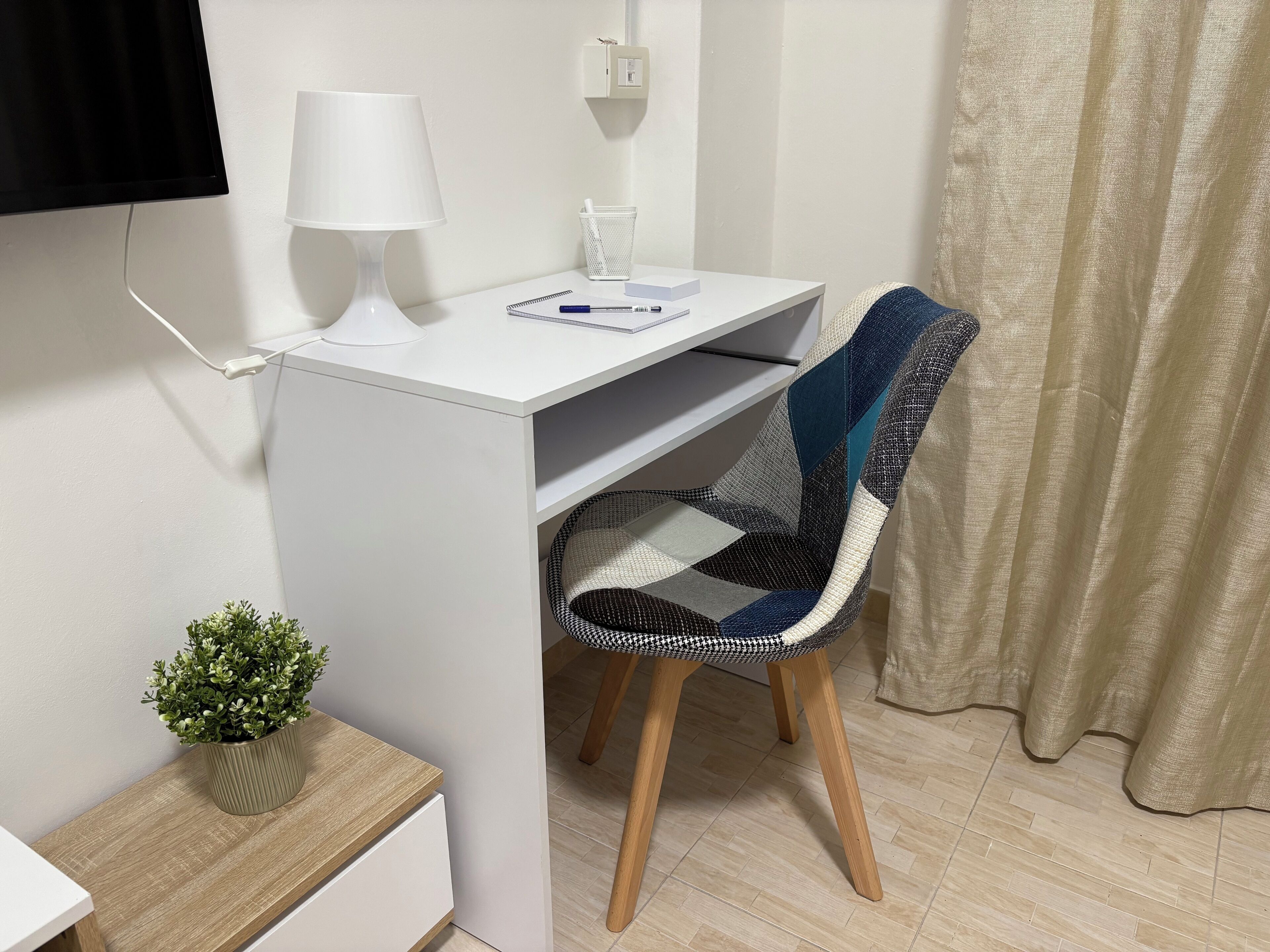 Una piccola scrivania bianca con una lampada, una sedia a quadretti e una pianta su una mensola in legno.