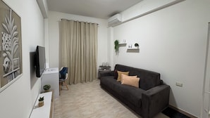 A modern living room with a grey sofa, a small side table, a flat-screen TV mounted on the wall, a desk with a chair, and a large window with curtains.