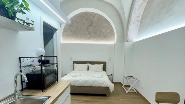 A compact bedroom with a bed, a small bedside table, a chair, and a kitchenette area with a sink and a microwave. - 1940 Apartment (Catania)