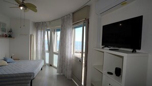 Ein Schlafzimmer mit einem Bett, einem Fernsehgerät auf einem Standfuß, einem Deckenventilator und einem Fenster mit Vorhängen.