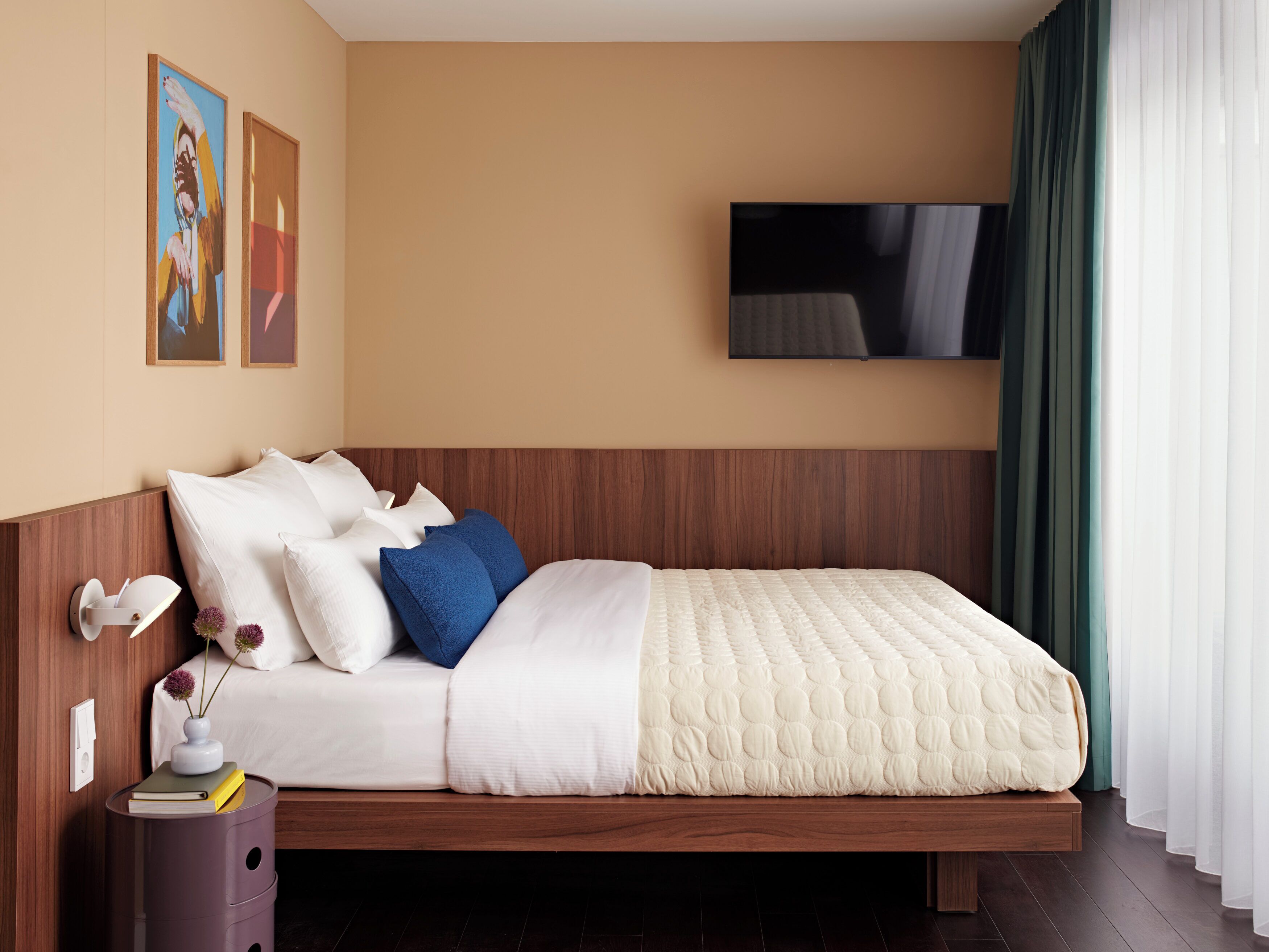 A modern hotel room with a wooden headboard, a flat-screen TV mounted on the wall, and a large window with sheer curtains.