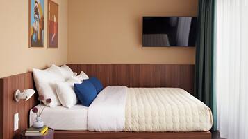 A modern hotel room with a wooden headboard, a flat-screen TV mounted on the wall, and a large window with sheer curtains.