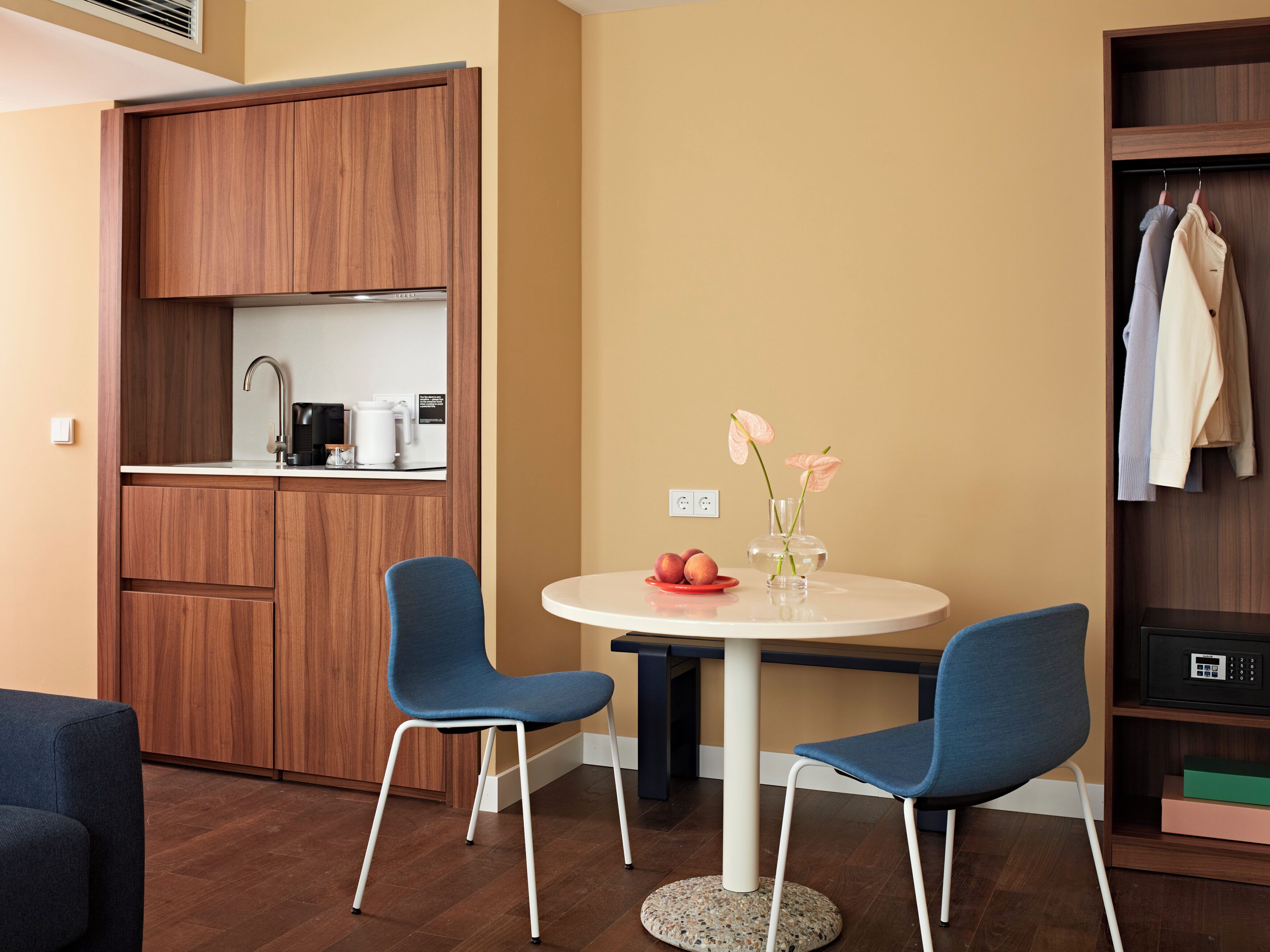 A compact living space with a kitchenette, two blue chairs, a small round table, and a vase with flowers.