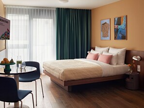 A hotel room with a bed, a desk with a chair, a table with fruits and a glass, and a window with curtains.