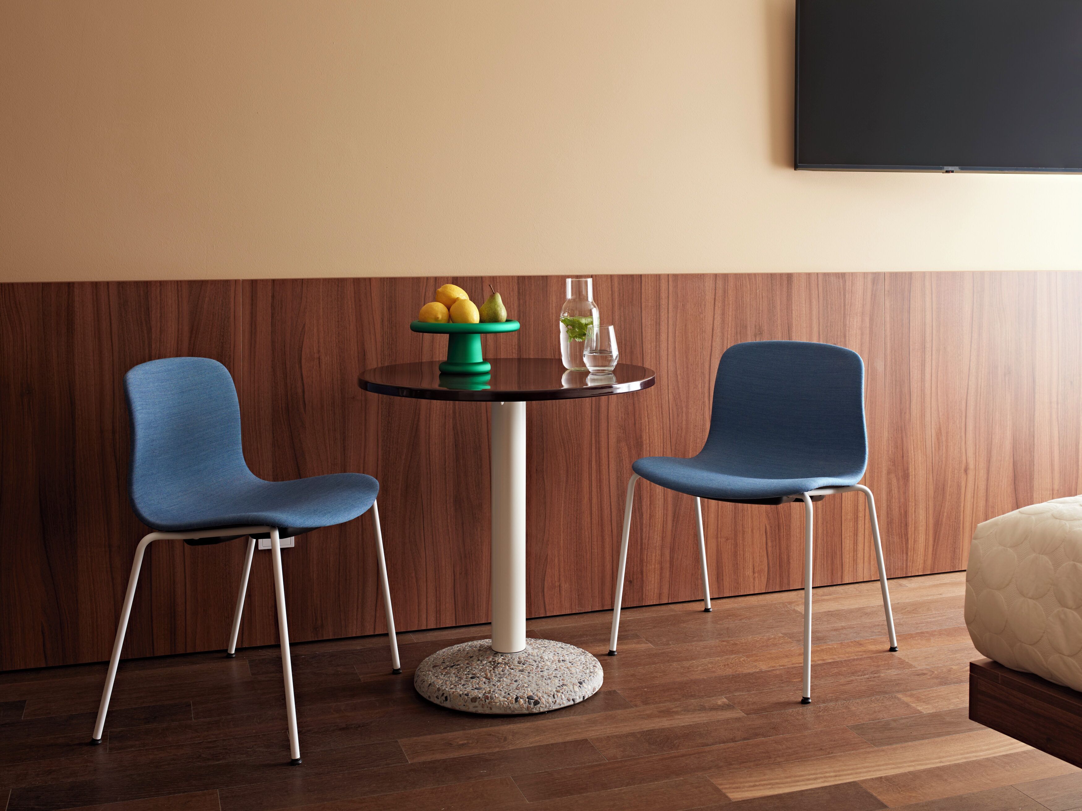 Une chambre moderne avec deux chaises bleues, une table ronde sur laquelle se trouve un panier de fruits, et un lit avec une tête de lit blanche.