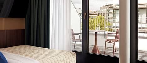 Un hotel moderno con cama, televisor montado en la pared, balcón con sillas y vistas a la ciudad.