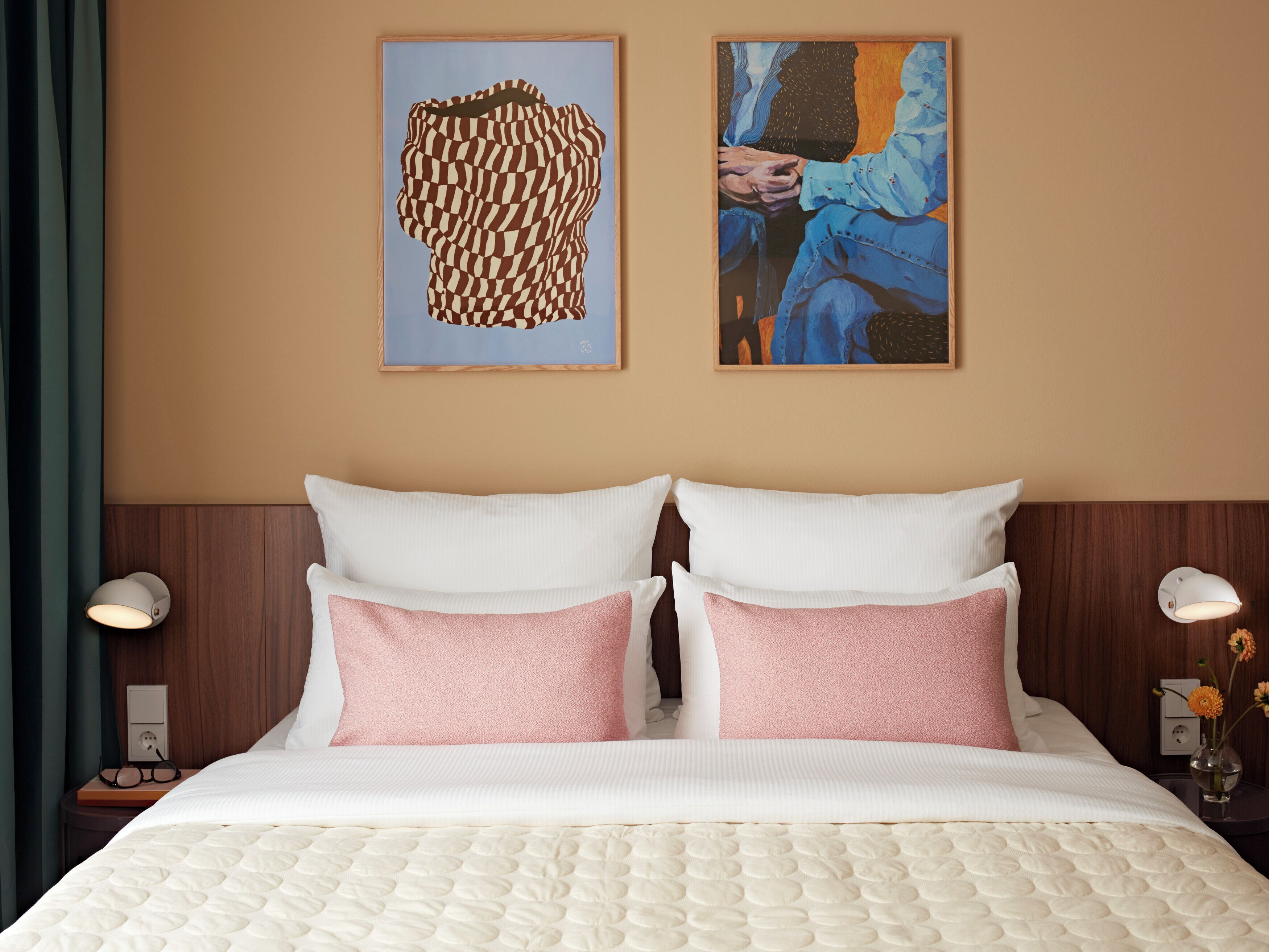 Hotelkamer met een bed, twee kussens, een hoofdbord, twee bedlampjes en twee kunstwerken in een lijst aan de muur.
