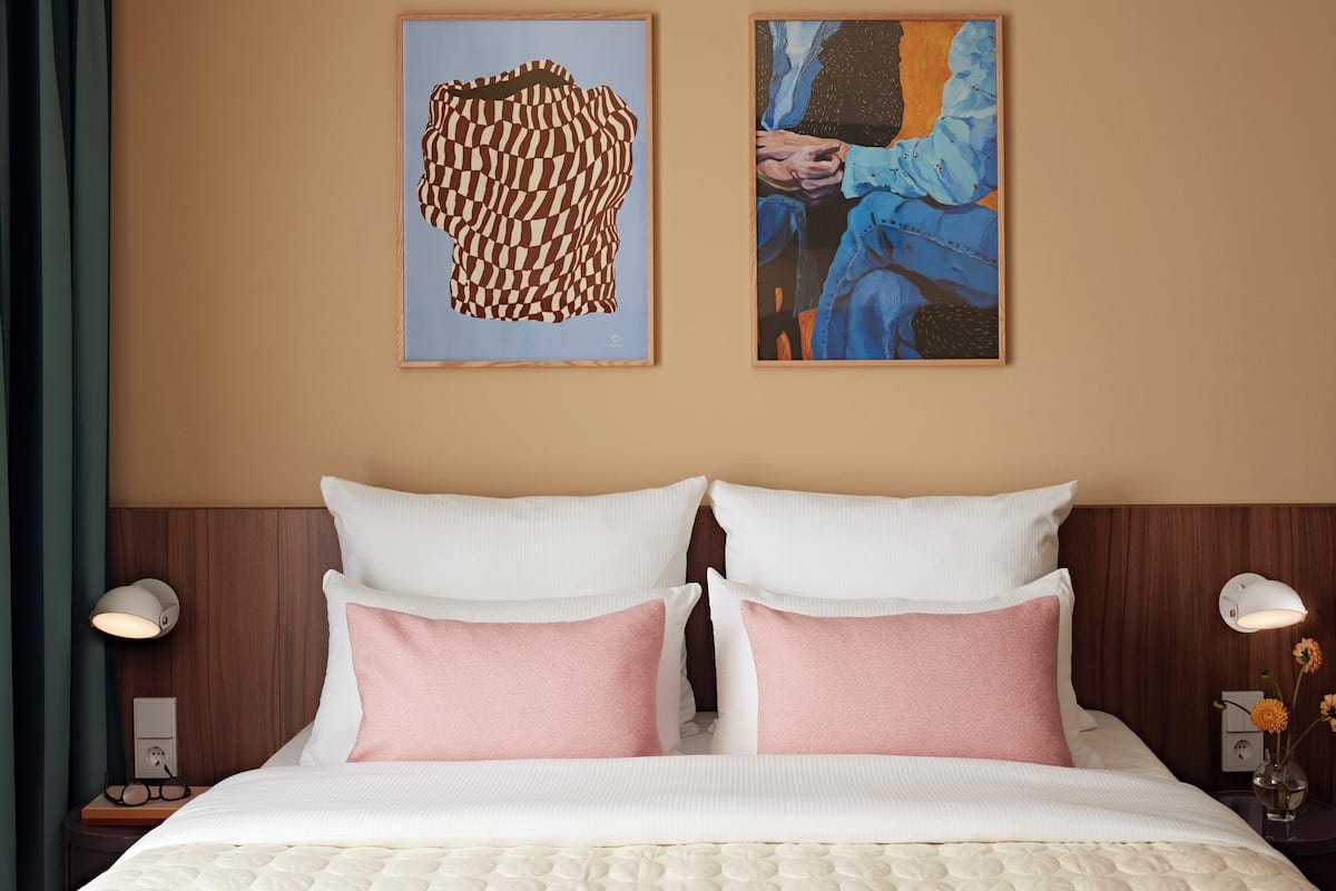 A hotel room with a bed, two pillows, a headboard, two bedside lamps, and two framed artworks on the wall.