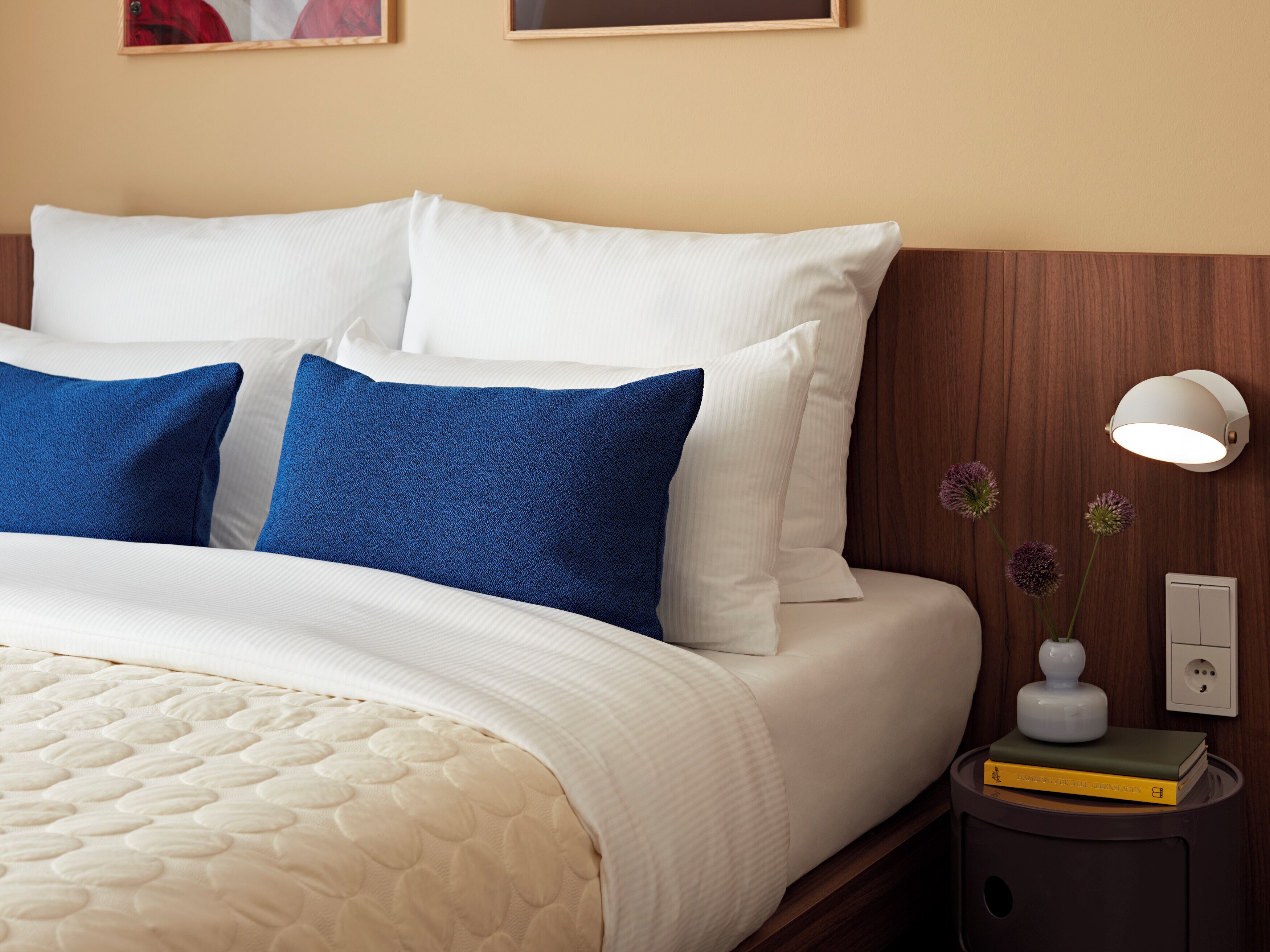 Een netjes opgemaakt bed met witte en blauwe kussens, een nachtkastje met een lamp en een kleine vaas met bloemen.