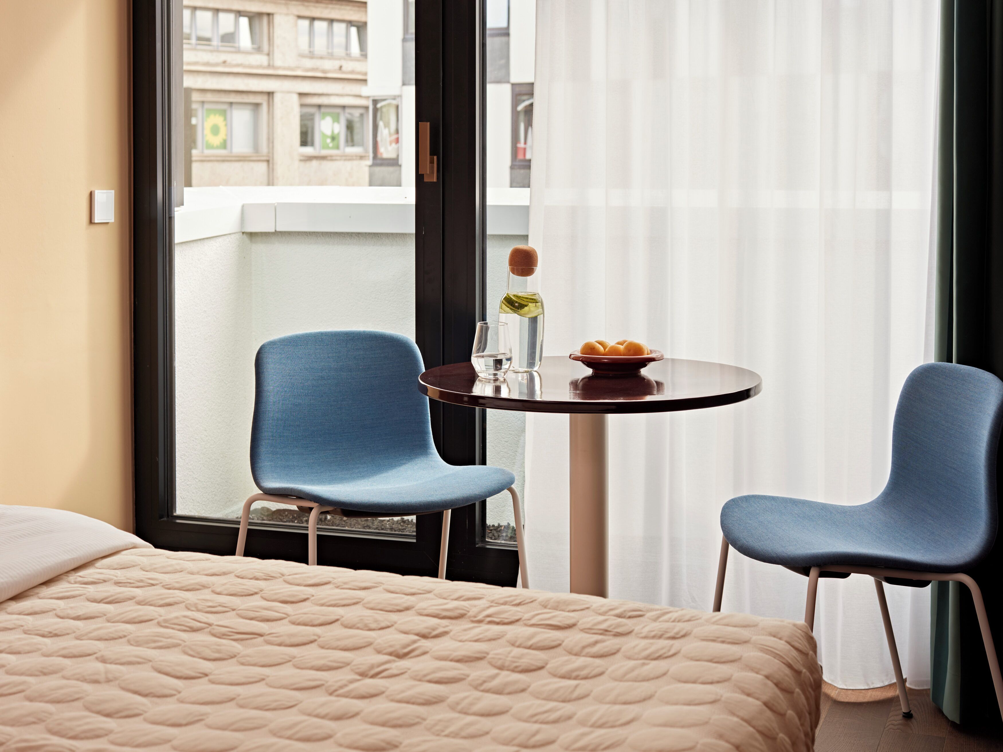 Ein Hotelzimmer mit einem Bett, zwei blauen Stühlen, einem kleinen runden Tisch mit einer Flasche und einer Schale mit Obst, sowie Blick auf Gebäude durch ein Fenster.