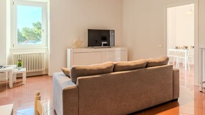 A modern living room with a beige sofa, a TV on a white cabinet, a radiator, and a window with a view of trees. - Nice apartment for 6 guests with WIFI, TV, balcony and pets allowed (Domaso)