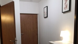 Un couloir avec une porte en bois, une commode blanche avec des tiroirs, un tableau encadré au mur et un luminaire au plafond.