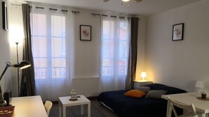Une pièce bien éclairée, avec un lit, un bureau, une chaise et une table. On y trouve des rideaux, un ventilateur de plafond et une fenêtre donnant sur un beau paysage.