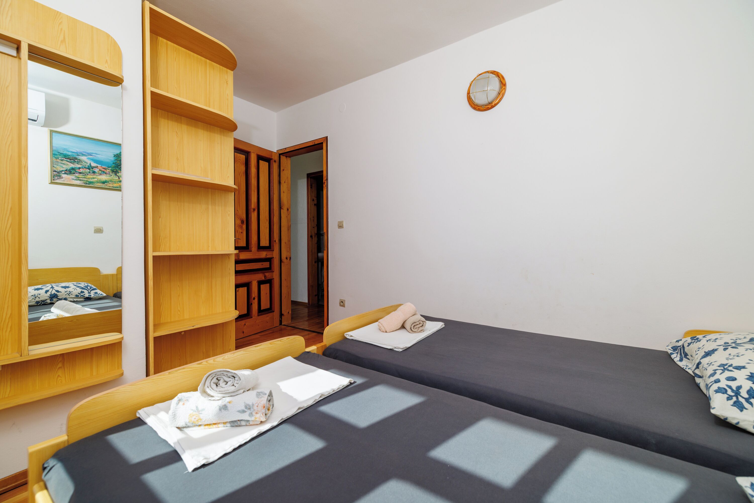 A room with two single beds, wooden furniture, a small clock on the wall, and a door leading to another room.