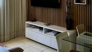 Un salon moderne avec un téléviseur à écran plat fixé à un mur en bois, une table à manger en verre avec des chaises, et un meuble de rangement blanc en dessous.