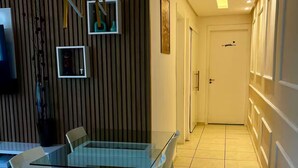 Un espace de salle à manger moderne avec une table en verre et des chaises blanches, un mur d’accent en bois et un couloir menant à une porte.