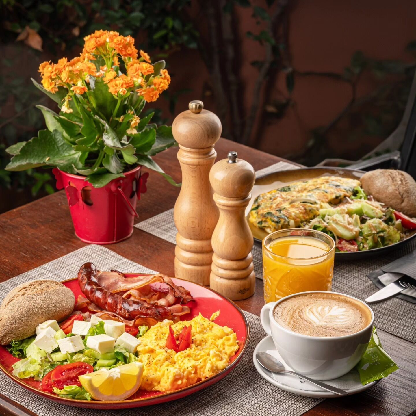 A breakfast setting with a plate of eggs, bacon, toast, and salad, a cup of coffee, a glass of orange juice, and a potted plant with orange flowers.