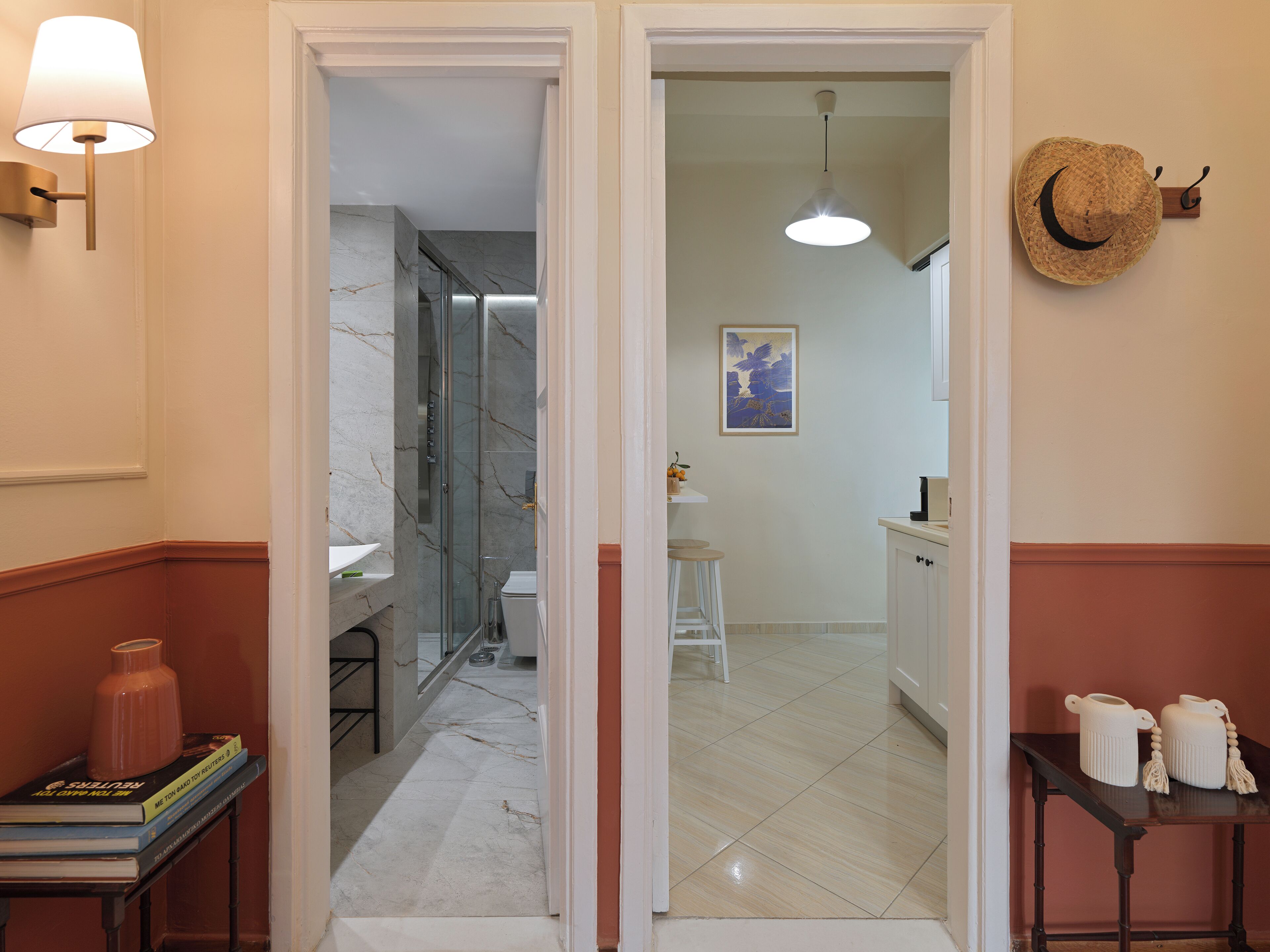 Un pasillo que conduce a un baño con ducha de vidrio, lavamanos e inodoro. A la izquierda hay una pequeña mesa con libros y un jarrón, y a la derecha una mesa con dos jarras.