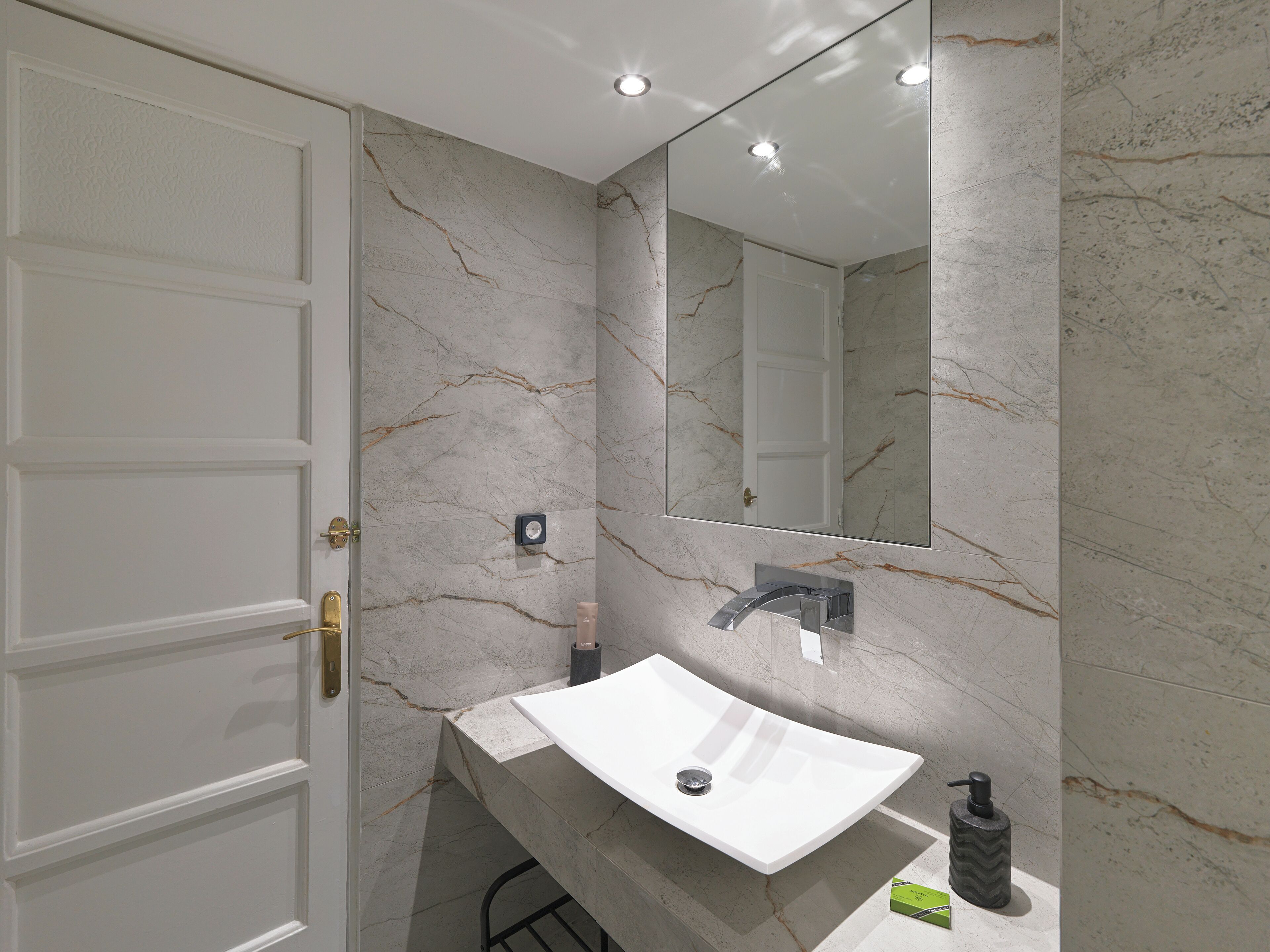 Un baño moderno con pared de mármol, un lavamanos blanco, un espejo y una puerta.