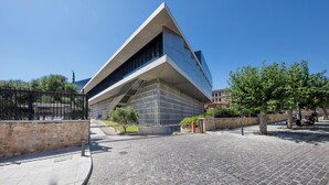 Un edificio moderno con un diseño angular distintivo, un área de entrada pavimentada y un cielo azul despejado.