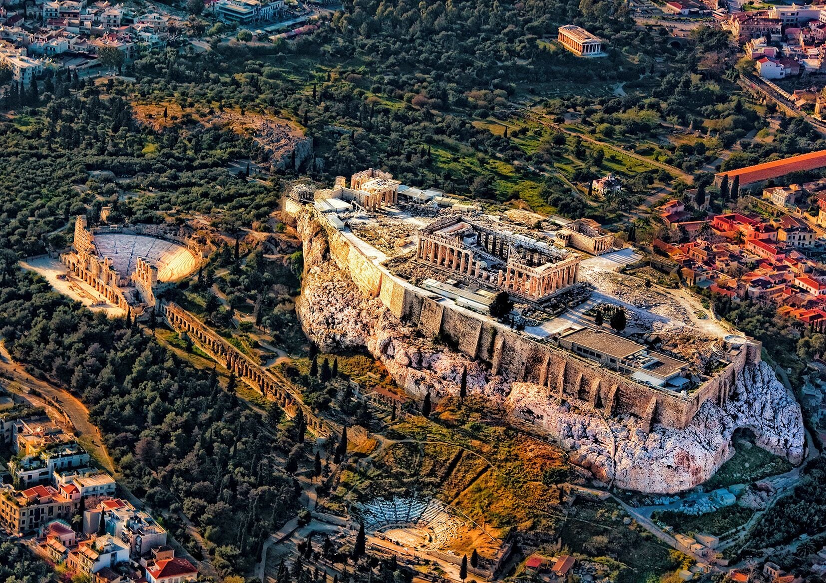 An aerial view of an ancient amphitheater with ruins, surrounded by dense vegetation and modern buildings.