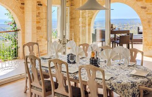 A dining area with a table set for six, surrounded by stone walls and large windows offering a view of the sea. - Le Mas d’Azur – Exceptional view & pool (Tourrettes-sur-Loup)