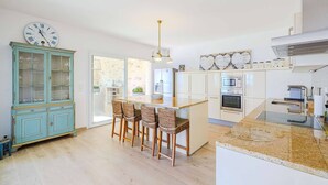 A modern kitchen with a central island, bar stools, a clock, and a cabinet with glass doors. - Le Mas d’Azur – Exceptional view & pool (Tourrettes-sur-Loup)