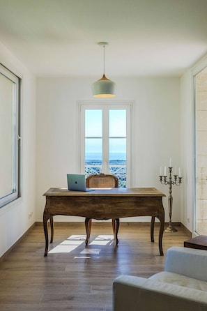 A room with a wooden table, a laptop, a wicker chair, a candelabra, and a view of the sea through a window. - Le Mas d’Azur – Exceptional view & pool (Tourrettes-sur-Loup)
