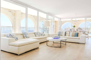 A modern living room with a large sofa set, a coffee table, and large windows offering a view of stone arches. - Le Mas d’Azur – Exceptional view & pool (Tourrettes-sur-Loup)