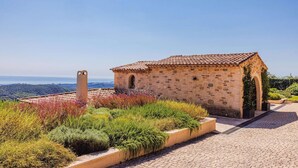 A stone building with a tiled roof, surrounded by a garden with various plants and flowers. - Le Mas d’Azur – Exceptional view & pool (Tourrettes-sur-Loup)