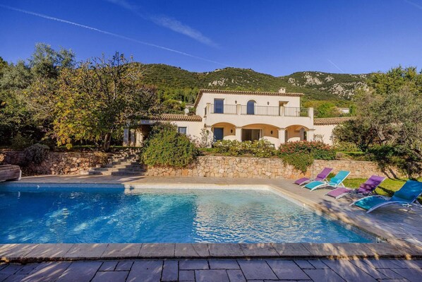 A pool with lounge chairs, a stone wall, and a building with a balcony.