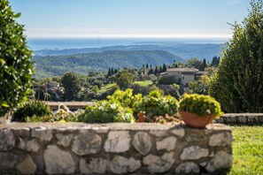 A stone wall with potted plants in the foreground, a stone building with a tiled roof in the middle ground, and a view of a coastal landscape with hills and the sea in the background.