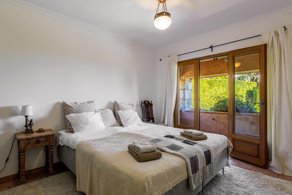 A bedroom with a bed, a nightstand, a lamp, a window with curtains, and a view of greenery outside.