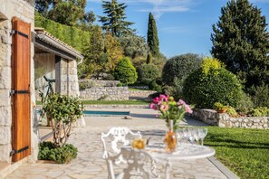 A stone building with a wooden door, a stone wall, and a garden with a pool, a table with chairs, and a vase of flowers.