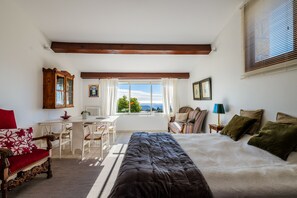A bedroom with a large bed, a desk, and a chair. There is a window with curtains, a lamp, and a view of the outdoors.