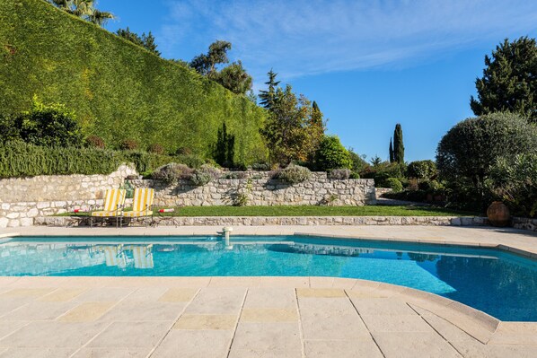 A swimming pool with stone tiles, two yellow-striped lounge chairs, and a stone wall with greenery.
