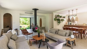 A modern living room with a large sofa, a dining table, and a fireplace. There is a view of the outdoors through a large window. - Villa Health – River & pool by Gorges du Loup (Tourrettes-sur-Loup)