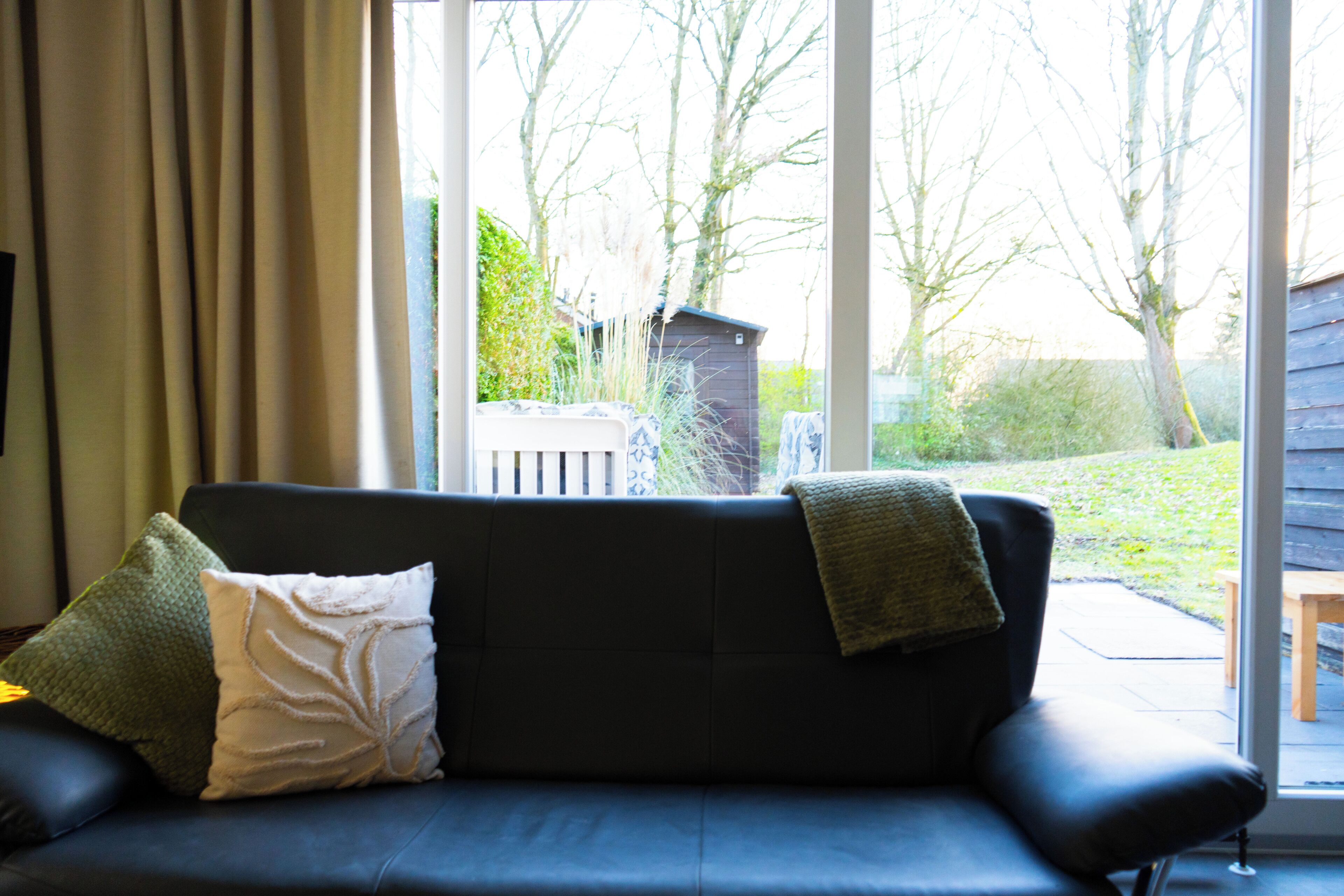 A dark-colored sofa with two cushions, a window with curtains, and a view of a wooden deck and trees outside.