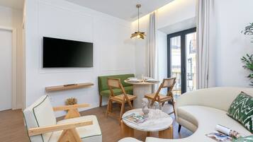 Una sala de estar moderna con un televisor de pantalla plana, sillas de madera, un sofá verde, una mesa de centro redonda y un amplio ventanal con cortinas.