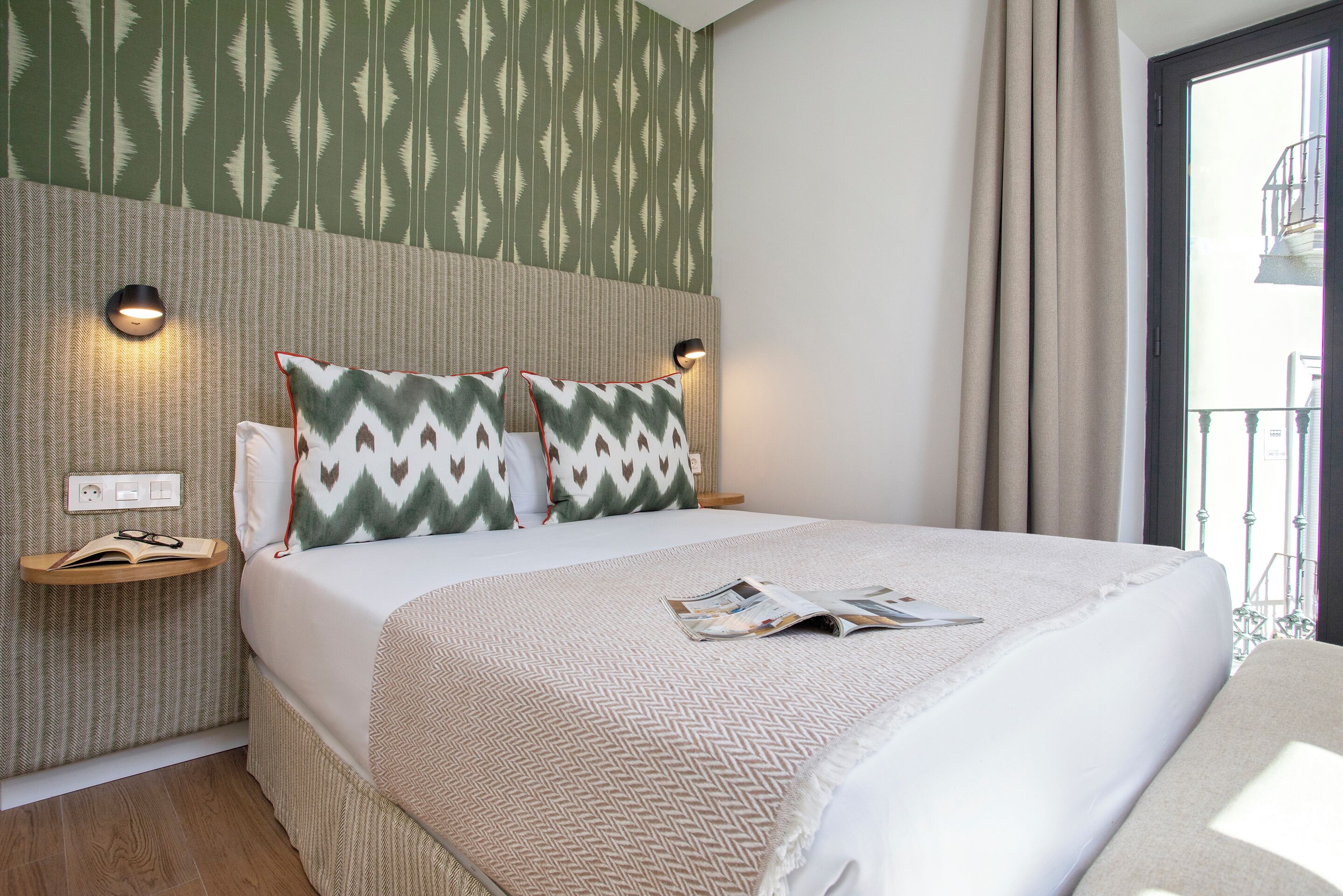A neatly made bed with patterned pillows, a bedside table with a lamp, and a magazine on the bed.