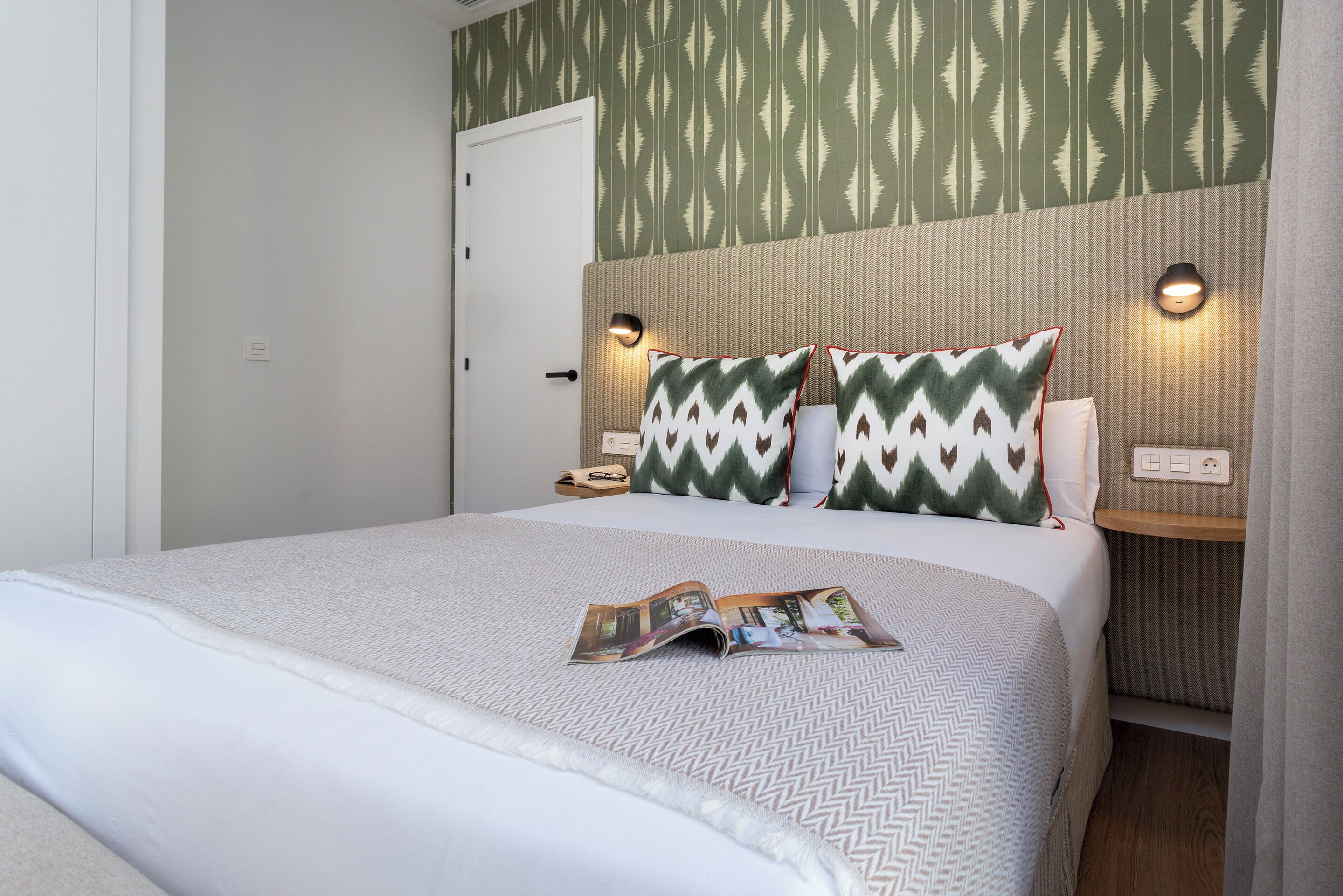 A neatly made bed with a magazine, two pillows, and a headboard with wall-mounted lights.