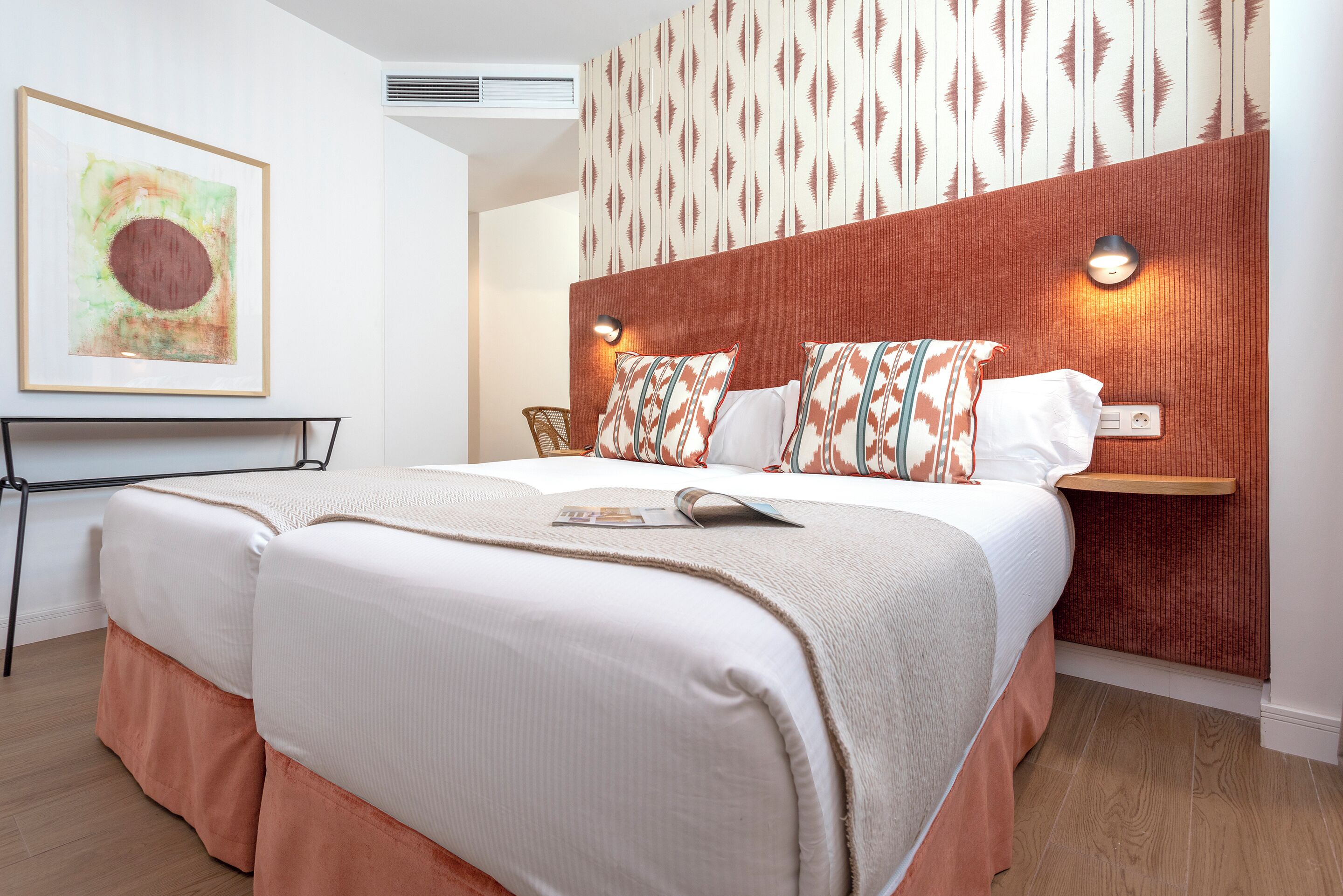 A modern hotel room with a large bed, a headboard with a patterned design, a wall-mounted light, and a framed artwork above the bed.