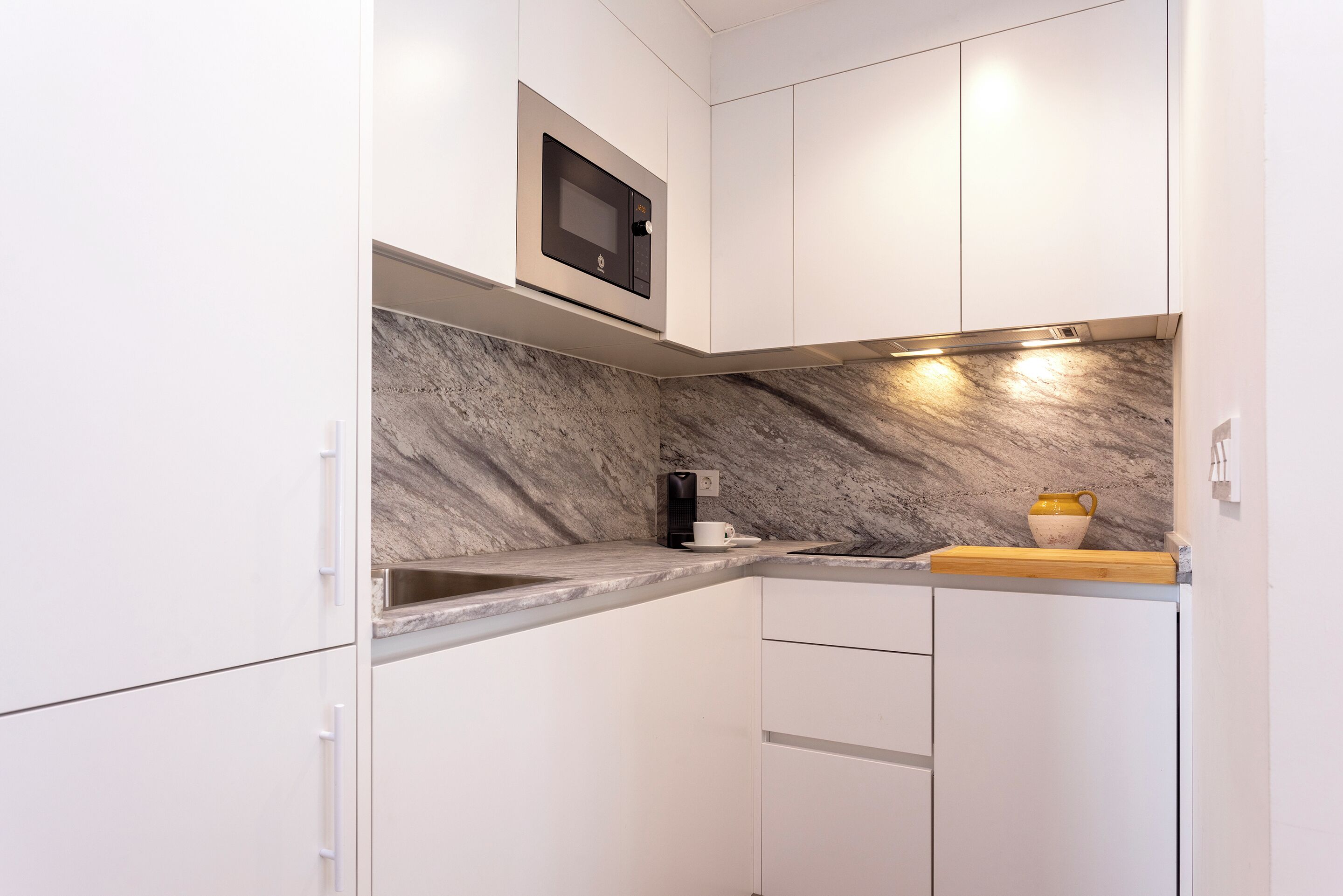Una cocina compacta con armarios blancos, microondas integrado, salpicadero de mármol y cafetera.
