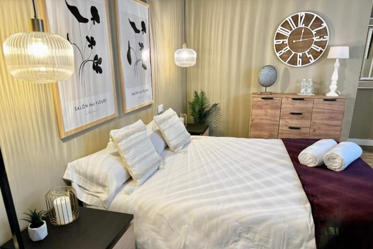 Una cama bien tendida con sábanas blancas, un aparador de madera con cajones, un reloj de pared redondo y cuadros en la pared.