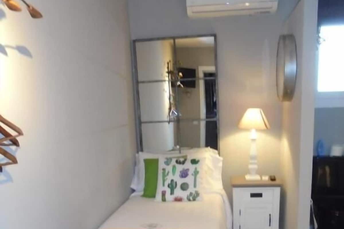 Un dormitorio con cama, mesita de noche, una silla, espejo y aire acondicionado.