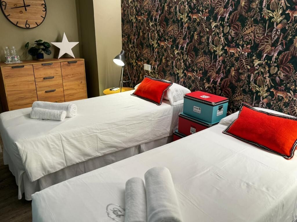 Dos camas con sábanas blancas y almohadas rojas, un aparador de madera, un reloj y un papel tapiz estampado.
