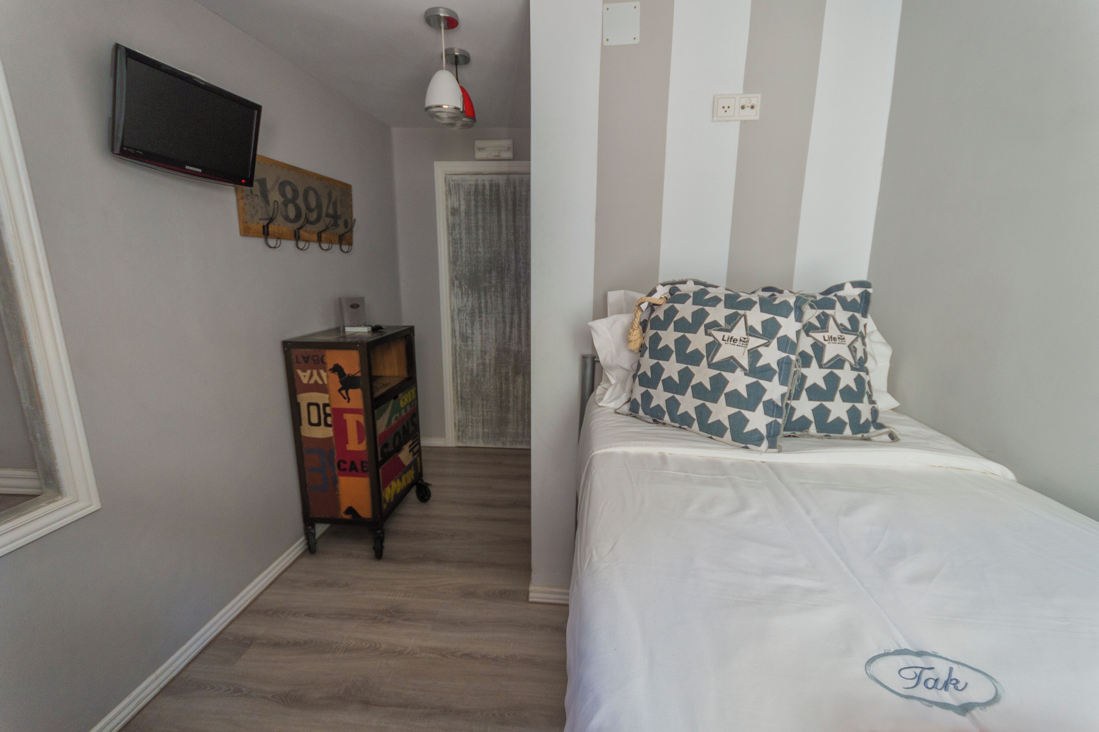 Un dormitorio con una cama, un televisor montado en la pared, un mueble pequeño y una puerta que da a otra habitación.