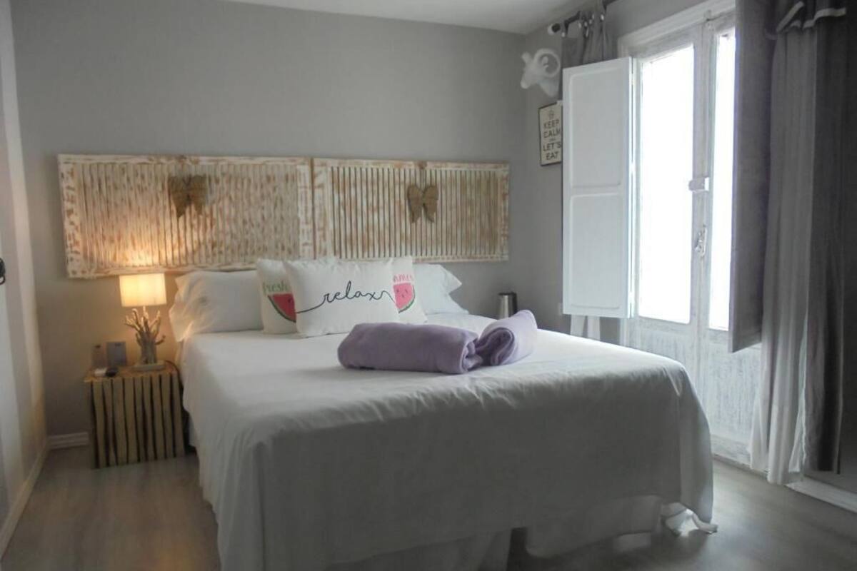 Una cama bien hecha con sábanas blancas y una toalla morada, una lámpara de noche y una obra de arte colgada en la pared sobre la cama.