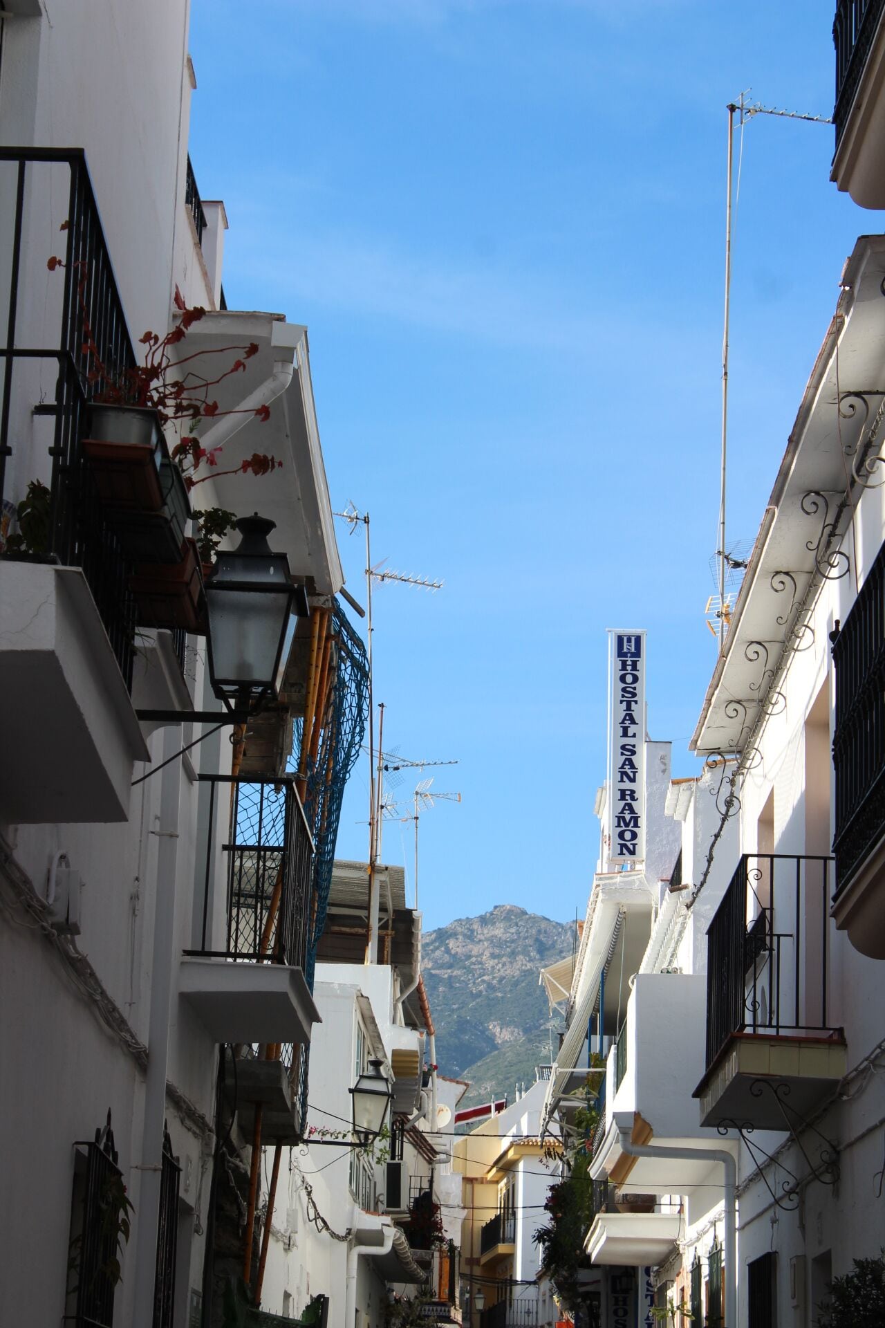 A narrow street with white buildings, balconies, and a mountain in the background.