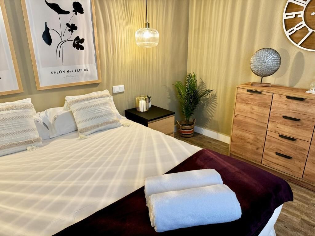 Ein ordentlich bezogenes Bett mit weißen Leinentüchern, einer dunkelroten Tagesdecke und zwei sauber gefalteten Handtüchern. Ein Holznachttisch mit einer kleinen Pflanze und einem dekorativen Gegenstand. Eine Holzkommode mit Schubladen befindet sich zur Rechten.